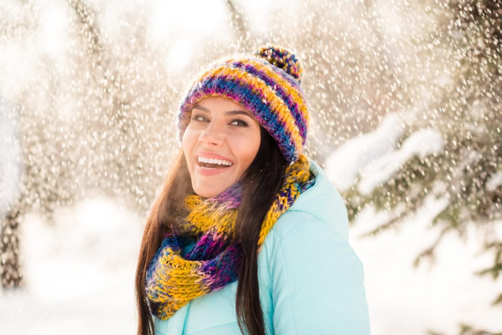 A woman wearing a colorful knit hat and scarf smiles outdoors as snow falls, reminding us of the importance of gum care during cold weather.